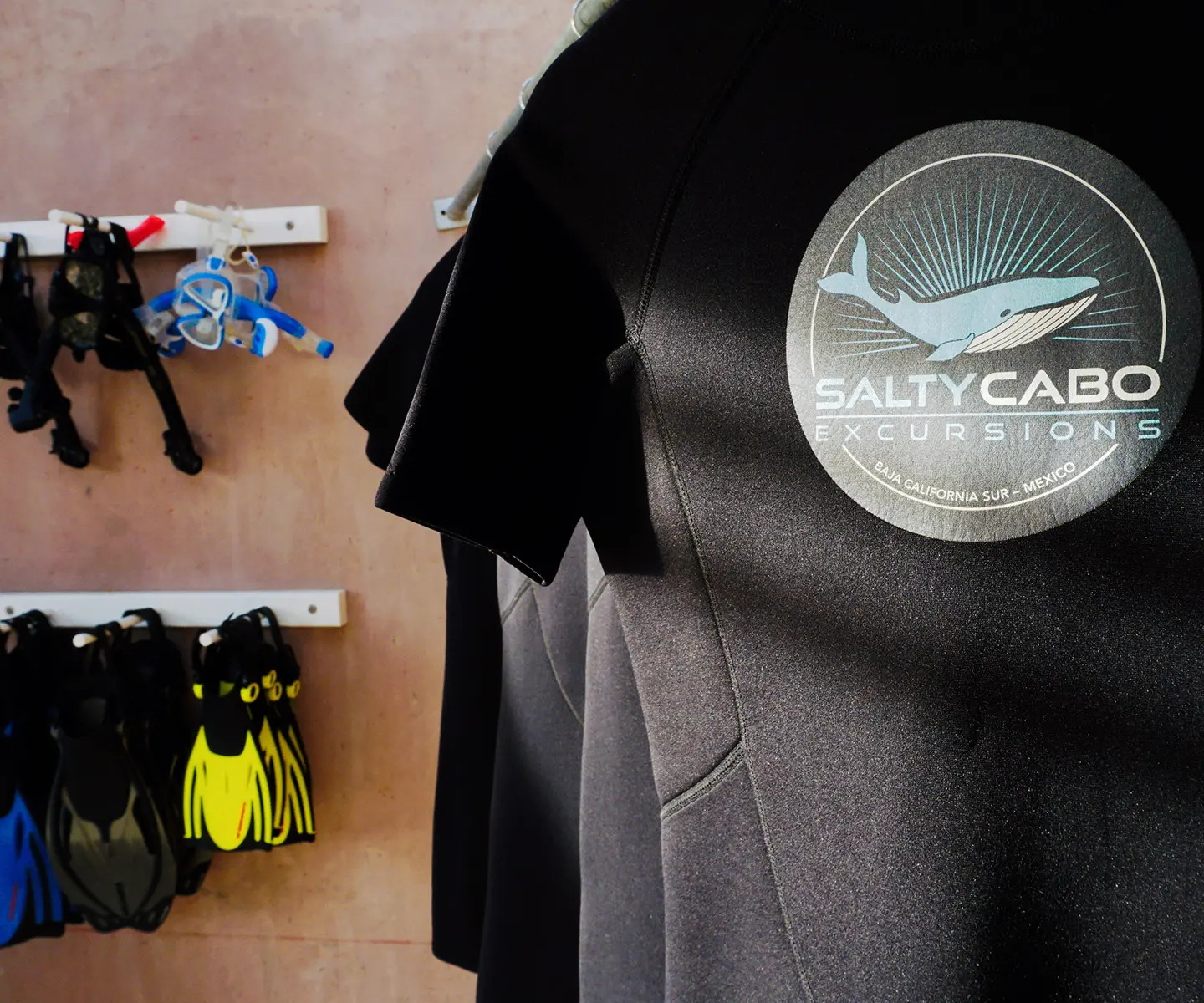 Close-up of Salty Cabo Excursions wetsuits with company logo, alongside snorkeling fins and equipment for tours in San José del Cabo.